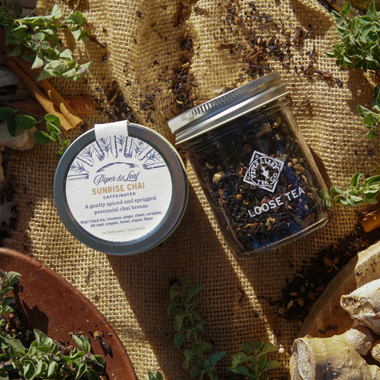 The Sunrise Chai Glass Jar of Loose Leaf Tea by Piper & Leaf Tea Co. sits on burlap with scattered tea leaves, cinnamon sticks, and ginger root, highlighting this vibrant spiced black tea blend. Contains 30 servings.