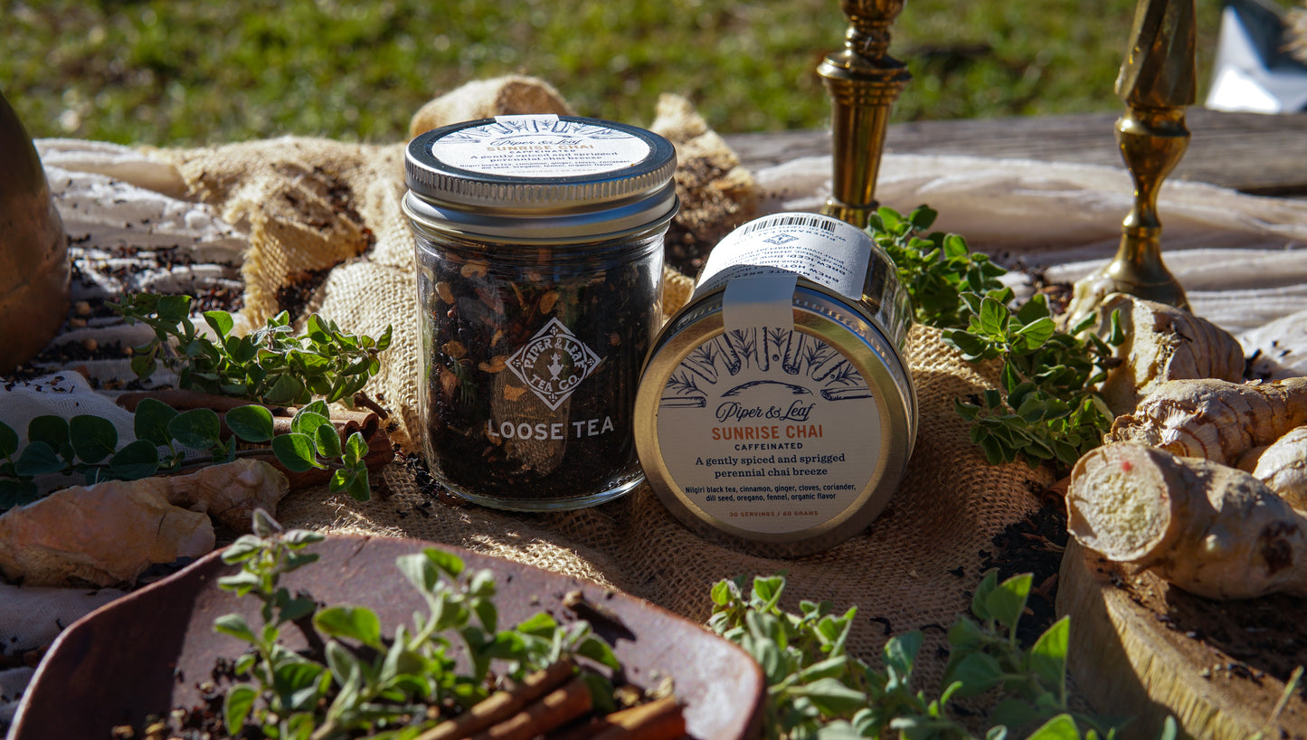 Two Piper & Leaf Tea Co. Sunrise Chai Glass Jars of Loose Leaf Tea (30 servings each) sit atop burlap, surrounded by fresh herbs, ginger, and cinnamon sticks on a wooden outdoor table.