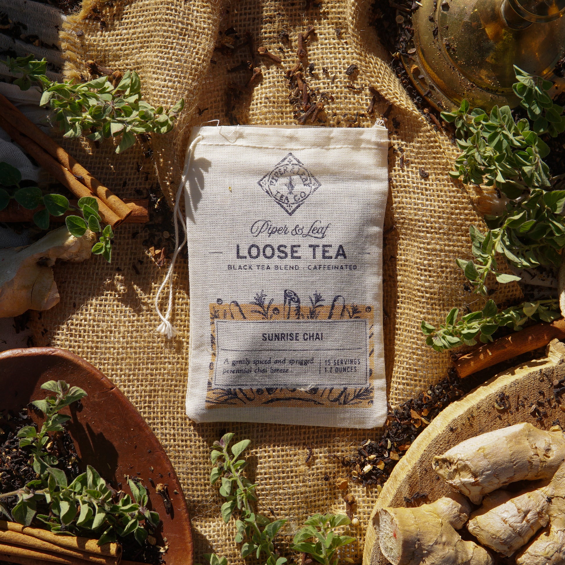 A muslin bag labeled "Piper & Leaf Tea Co. Sunrise Chai Loose Leaf Tea - 15 Servings" sits on burlap, surrounded by ginger, cinnamon sticks, herbs, and loose tea—ideal for spiced black tea lovers.