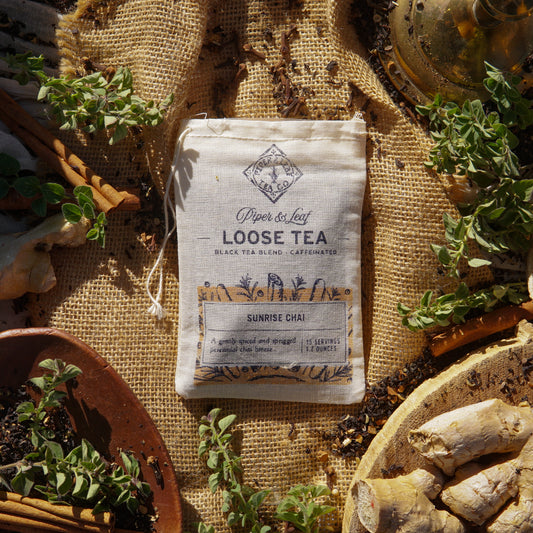 A muslin bag labeled "Piper & Leaf Tea Co. Sunrise Chai Loose Leaf Tea - 15 Servings" sits on burlap, surrounded by ginger, cinnamon sticks, herbs, and loose tea—ideal for spiced black tea lovers.