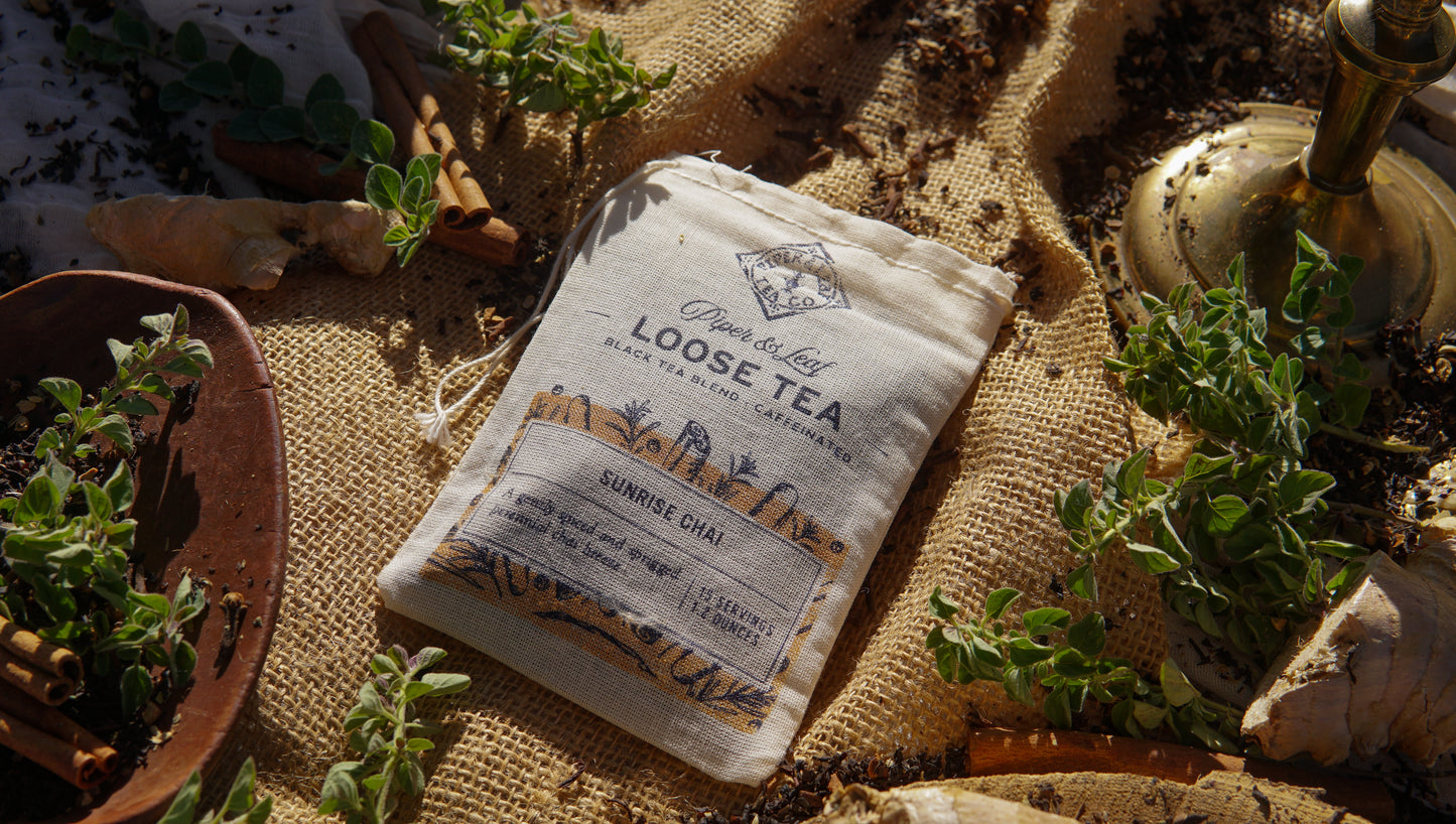 A muslin bag labeled "Sunrise Chai Loose Leaf Tea - 15 Servings" by Piper & Leaf Tea Co. rests on burlap amid herbs, ginger roots, and a brass lamp in sunlight, evoking the rich aroma of chai tea.