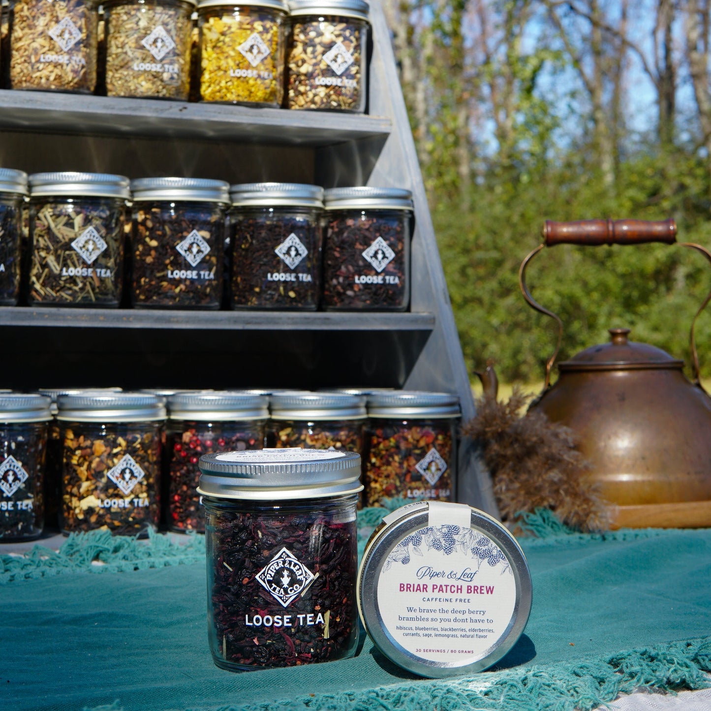 Jars of loose leaf tea from Piper & Leaf Tea Co. are displayed outdoors, featuring the "Briar Patch Brew" Glass Jar (30 Servings), a caffeine-free berry tea, alongside an old-fashioned kettle on a teal cloth in front.