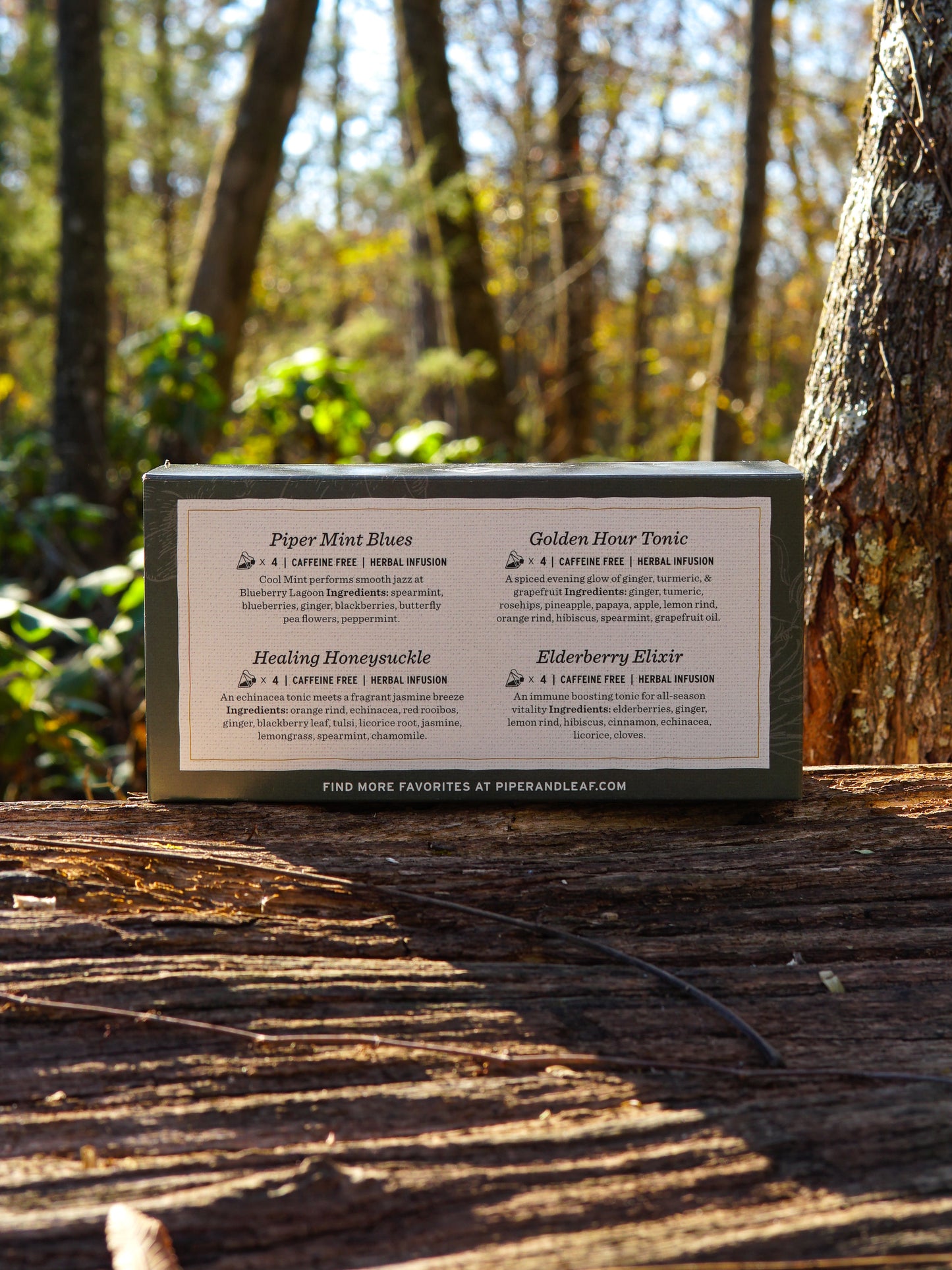 A Piper & Leaf Tea Co. Wellness Tea Sampler—Box of 16 Tea Bags—rests on a wooden log outdoors among trees and sunlight, ideal for anyone seeking a soothing herbal infusion tea experience.