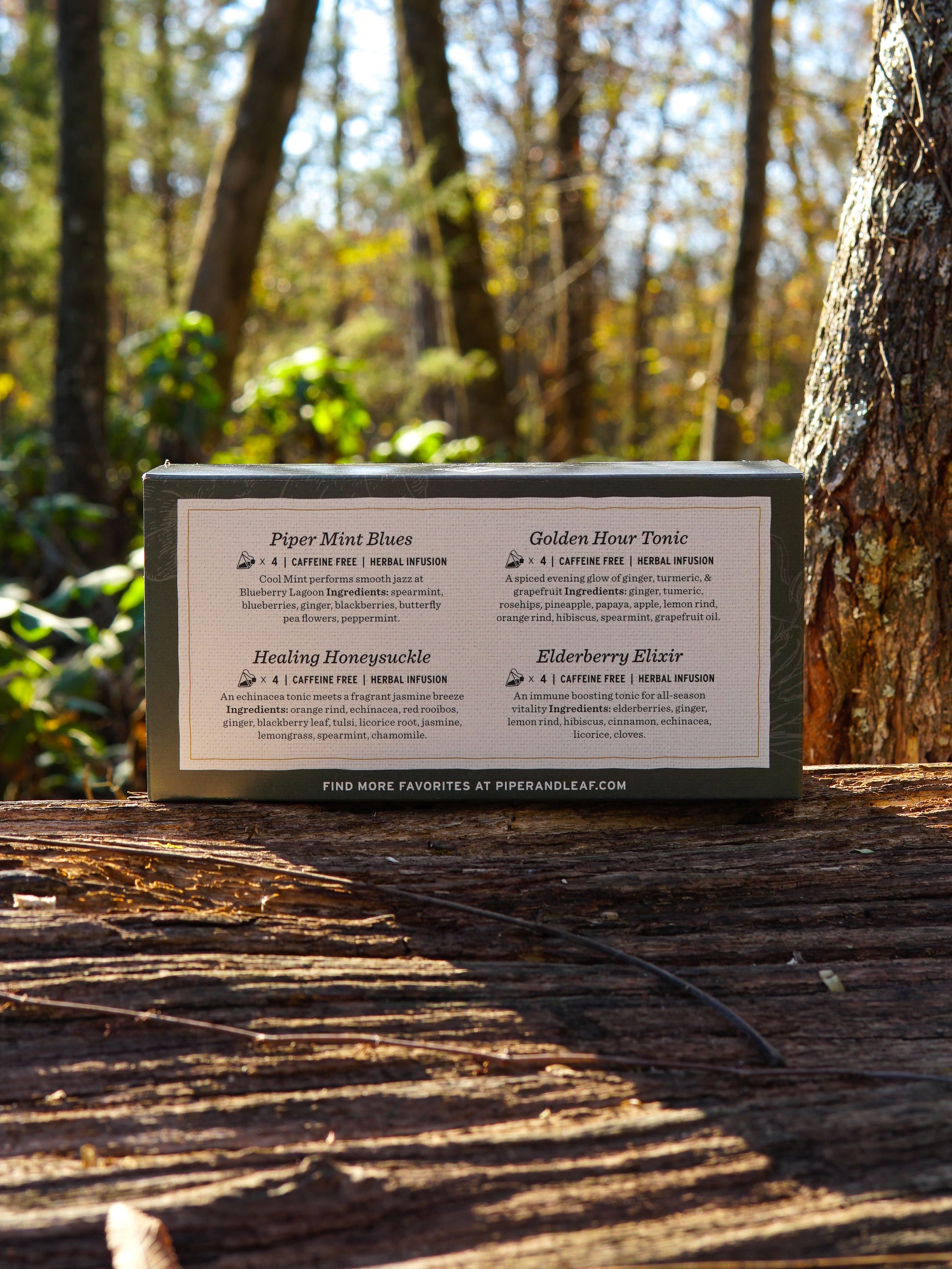 A Piper & Leaf Tea Co. Wellness Tea Sampler—Box of 16 Tea Bags—rests on a wooden log outdoors among trees and sunlight, ideal for anyone seeking a soothing herbal infusion tea experience.