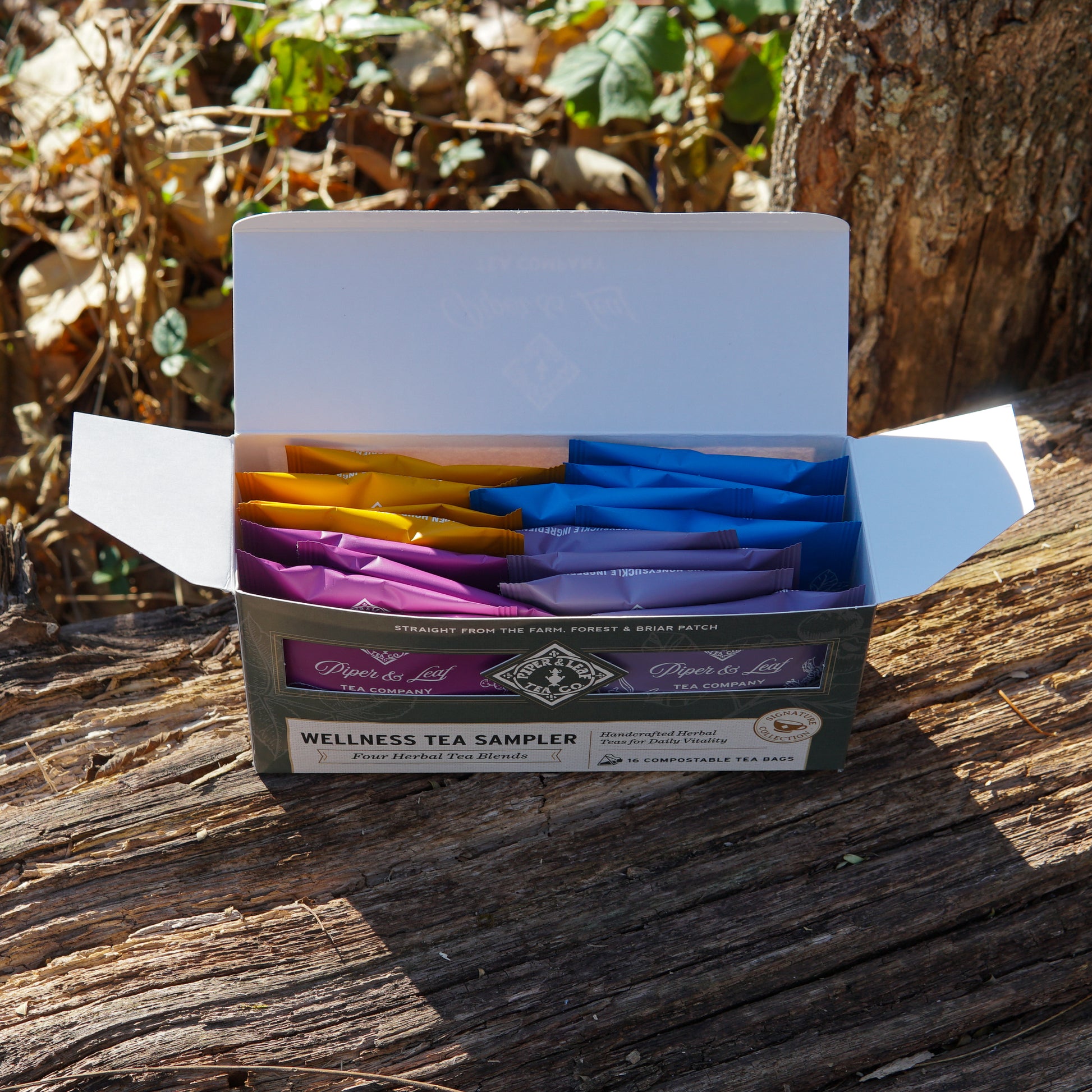 A box of Piper & Leaf Tea Co. Wellness Tea Sampler—16 caffeine-free tea bags—sits on a wooden surface outdoors, with tree bark and dried leaves in the background.