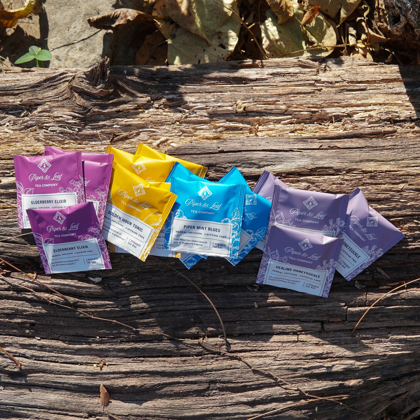 The Wellness Tea Sampler by Piper & Leaf Tea Co.—a box of 16 herbal infusion tea bags—is displayed on a weathered wooden surface with dried leaves in the background.