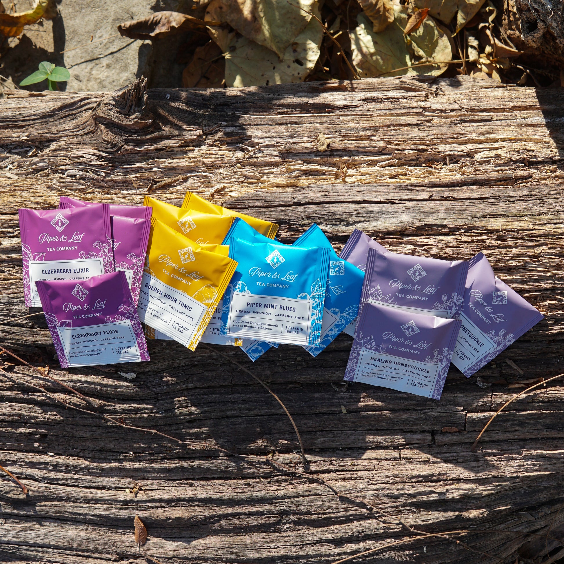 The Wellness Tea Sampler by Piper & Leaf Tea Co.—a box of 16 herbal infusion tea bags—is displayed on a weathered wooden surface with dried leaves in the background.