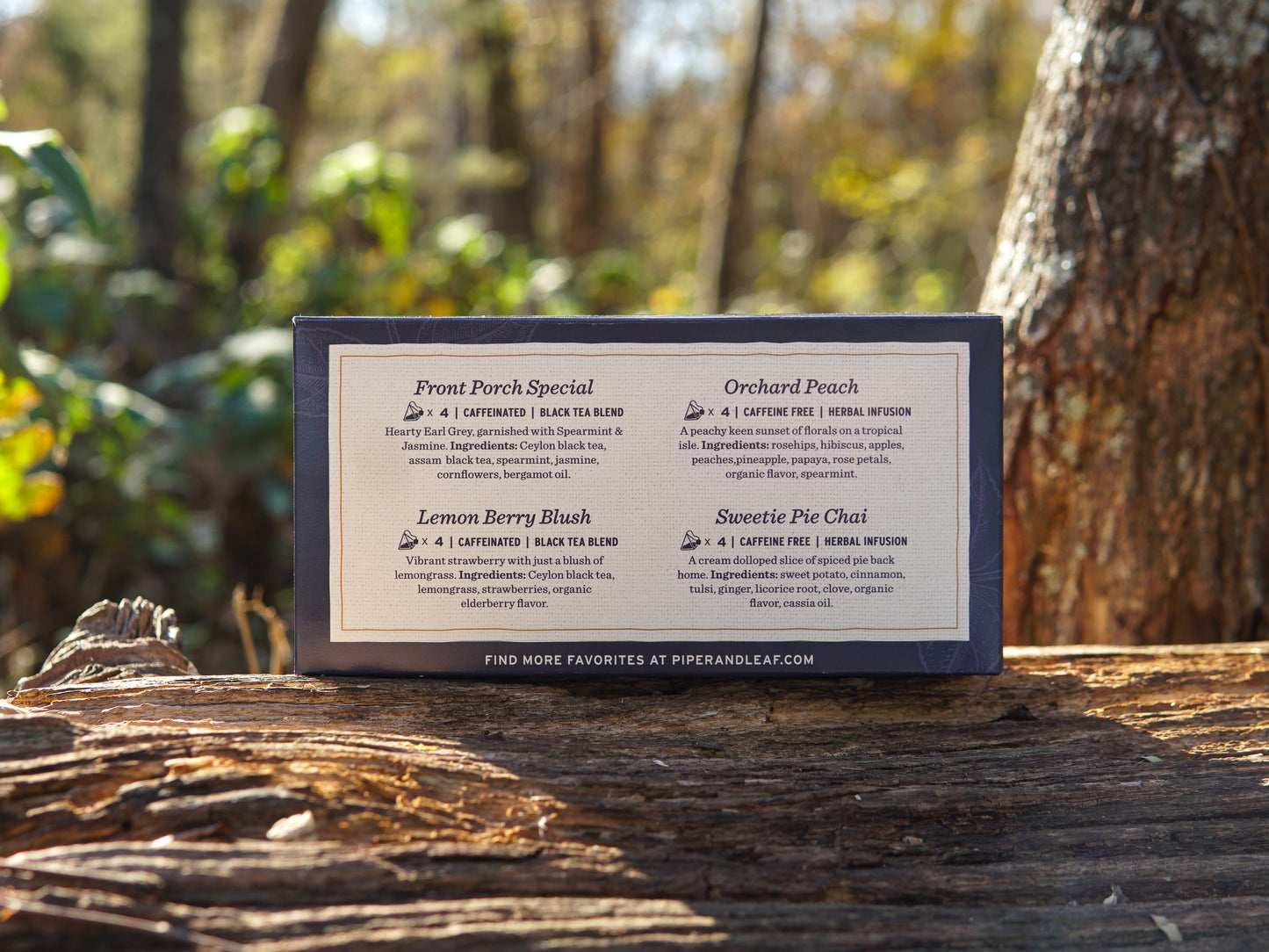 A Bestselling Tea Sampler Box of 16 Tea Bags by Piper & Leaf Tea Co. rests on a sunlit tree trunk, displaying four blends—Front Porch Special, Orchard Peach, Lemon Berry Blush (herbal), and Sweetie Pie Chai—against a forest backdrop.
