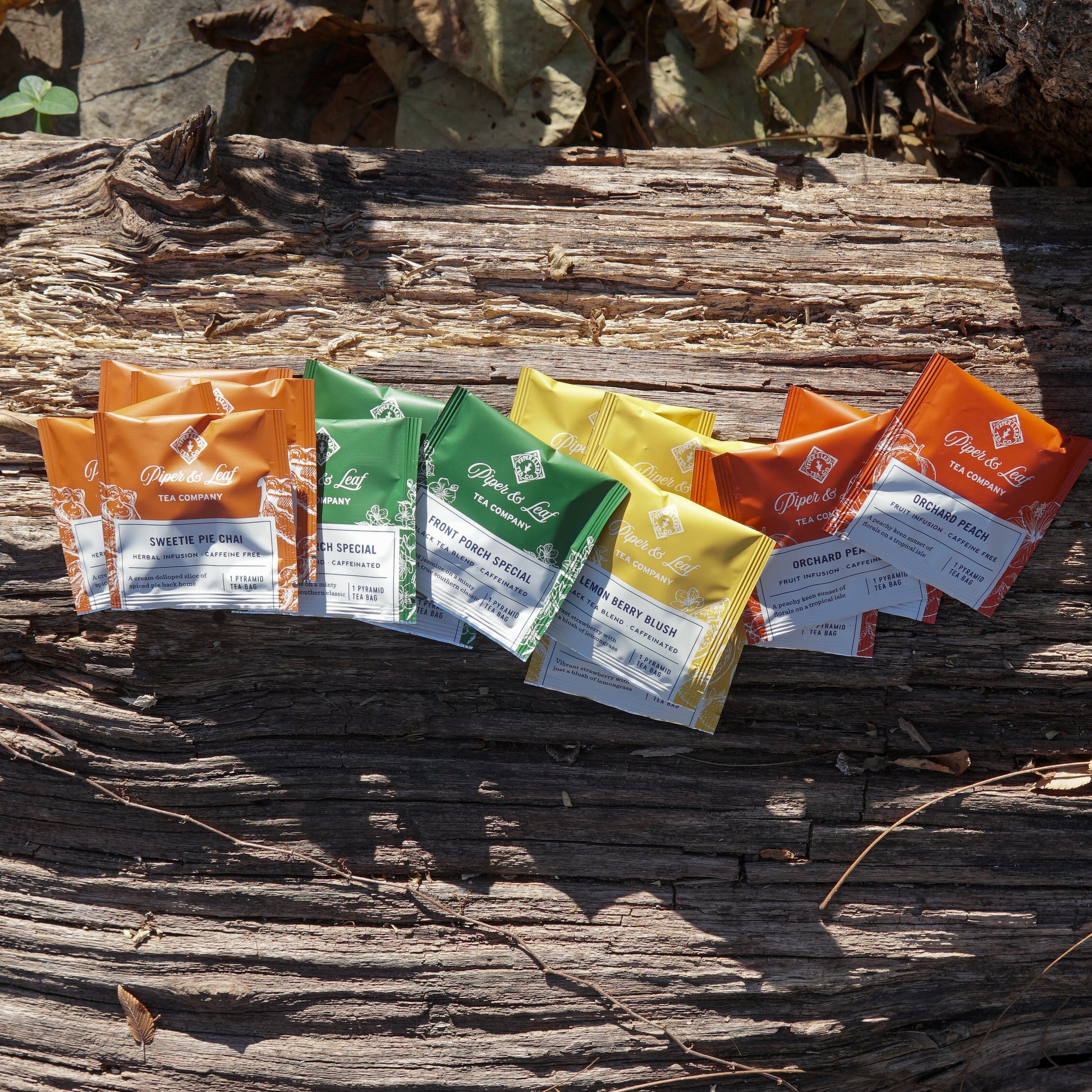 Piper & Leaf Tea Co.'s Bestselling Tea Sampler (box of 16 bags) in orange, green, and yellow wrappers is displayed on a weathered wooden surface with dry leaves, featuring both black tea blends and caffeine-free herbal infusions.