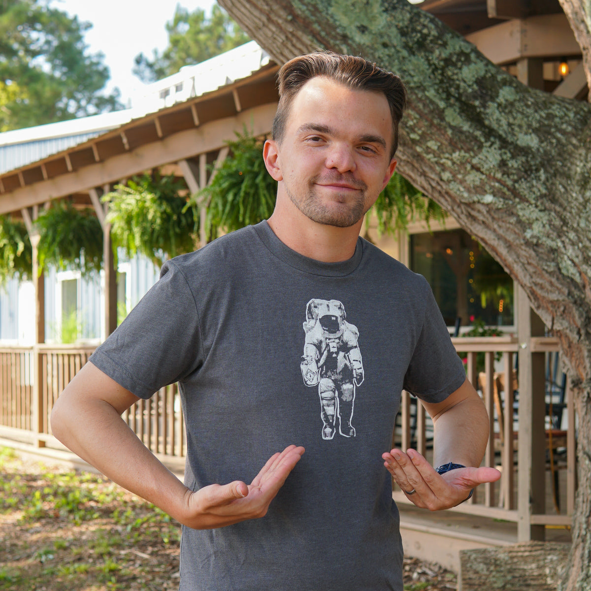 A person stands outside, pointing at a graphic of an astronaut on their grey Space Man Classic Tee-Shirt by Piper & Leaf Tea Co. Trees, ferns, and a wooden building appear in the background, reflecting Rocket City’s passion for all things cosmic.