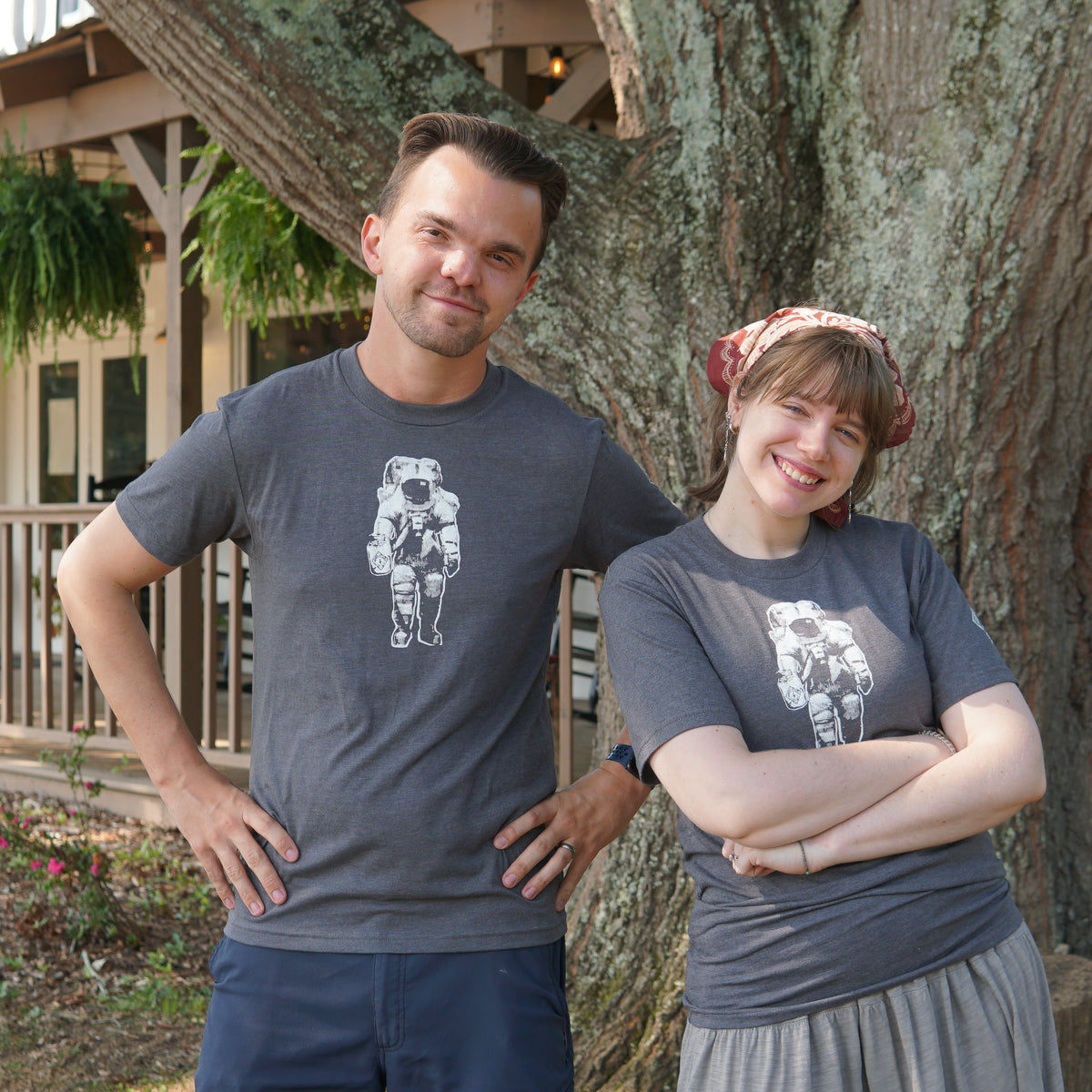 Two people stand in front of a tree in Huntsville, both wearing Piper & Leaf Tea Co.’s Space Man Classic Tee-Shirt in grey. They are smiling and posing with arms akimbo, proudly showing off their Rocket City style.