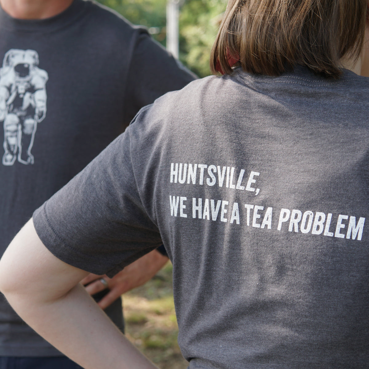 Two people wear dark gray T-shirts from Piper & Leaf Tea Co.; one shirt showcases the playful phrase “HUNTSVILLE, WE HAVE A TEA PROBLEM” in white, while the other is the Space Man Classic Tee-Shirt - Grey, featuring a space-themed graphic of an astronaut.