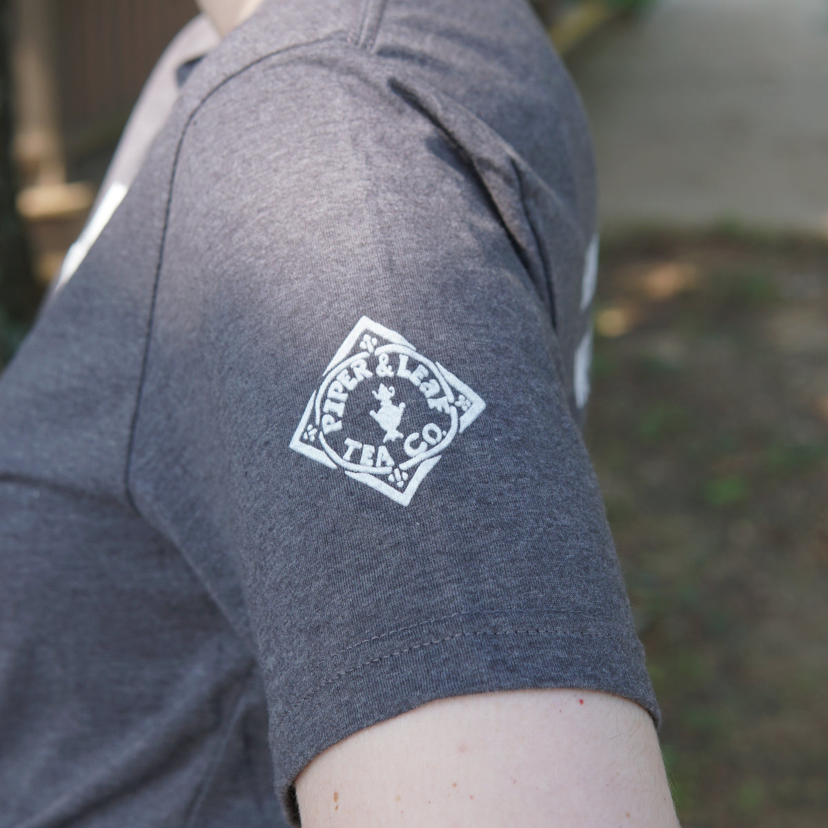 A person wearing the Space Man Classic Tee-Shirt in grey by Piper & Leaf Tea Co., featuring a white logo on the sleeve, photographed outdoors in Rocket City, Huntsville.