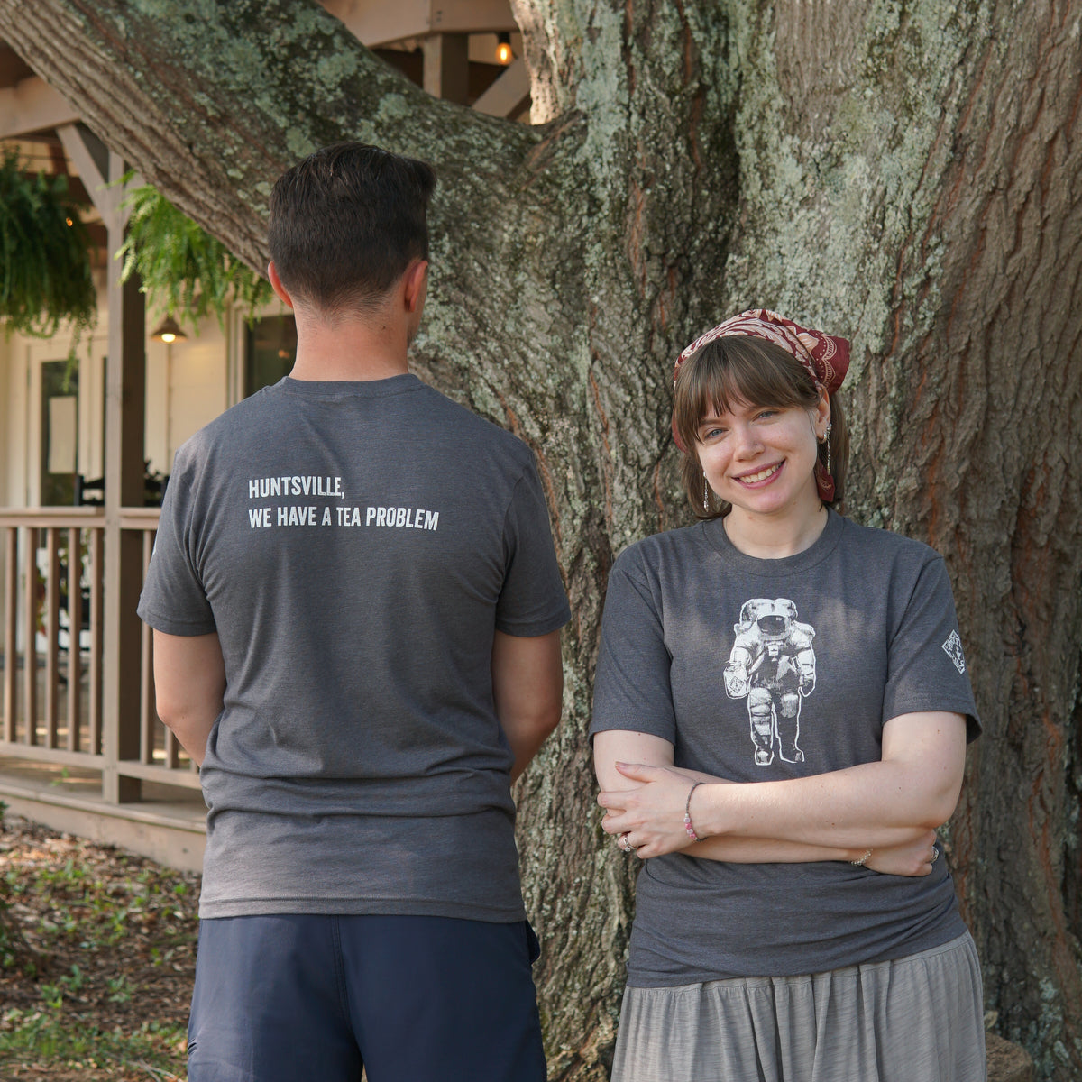 Two people stand by a tree; one faces away, wearing a shirt that says “Huntsville We Have a Tea Problem” by Piper & Leaf Tea Co., while the other faces forward in the Space Man Classic Tee-Shirt - Grey, which features an astronaut graphic—a perfect nod to Rocket City’s cosmic charm.