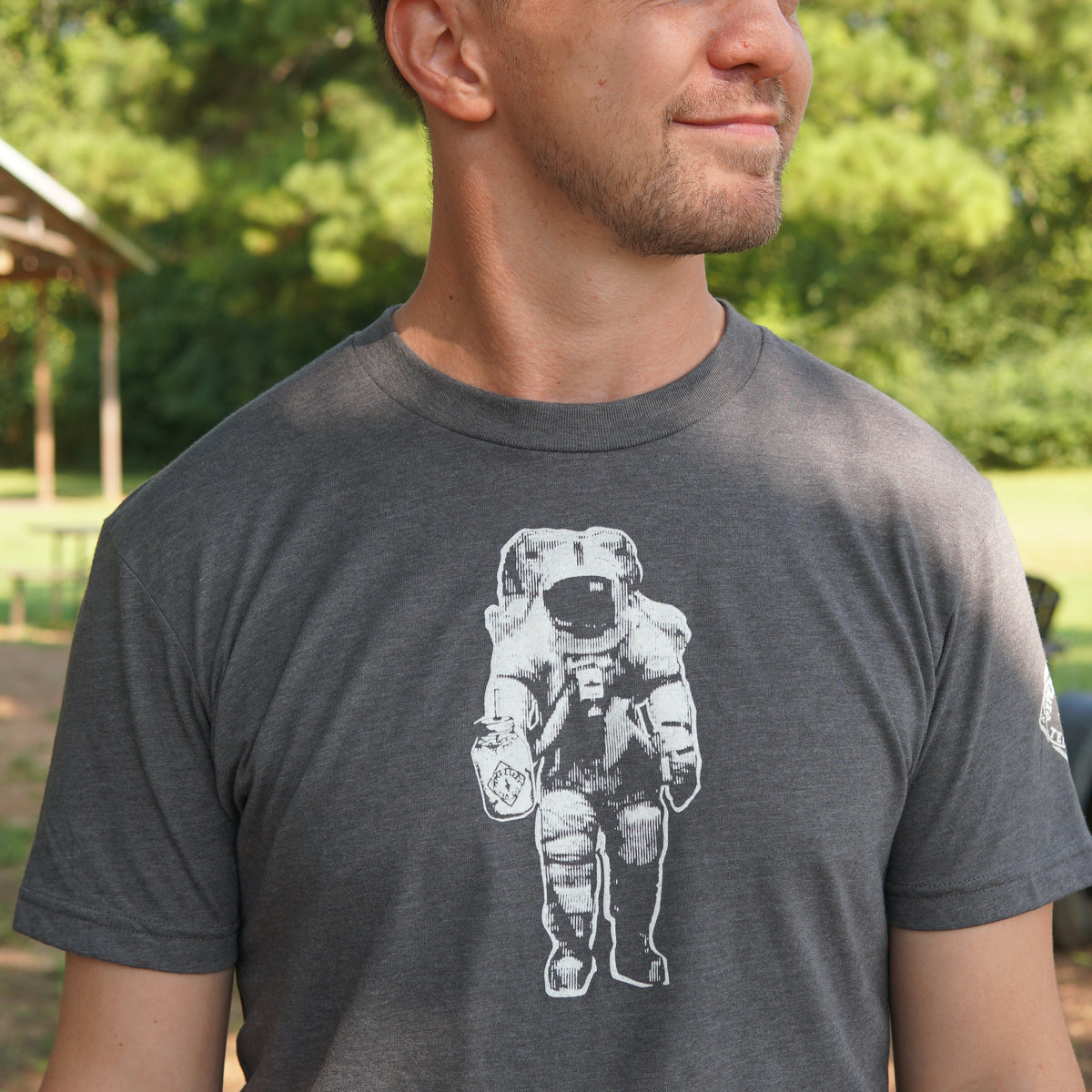 Wearing the Piper & Leaf Tea Co. Space Man Classic Tee-Shirt in grey, featuring a bold white astronaut graphic, a person stands outdoors among trees—an ideal representation of Rocket City, Huntsville.
