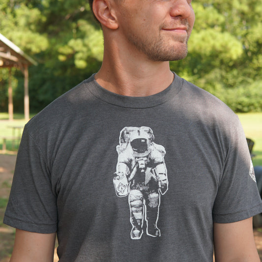 Wearing the Piper & Leaf Tea Co. Space Man Classic Tee-Shirt in grey, featuring a bold white astronaut graphic, a person stands outdoors among trees—an ideal representation of Rocket City, Huntsville.