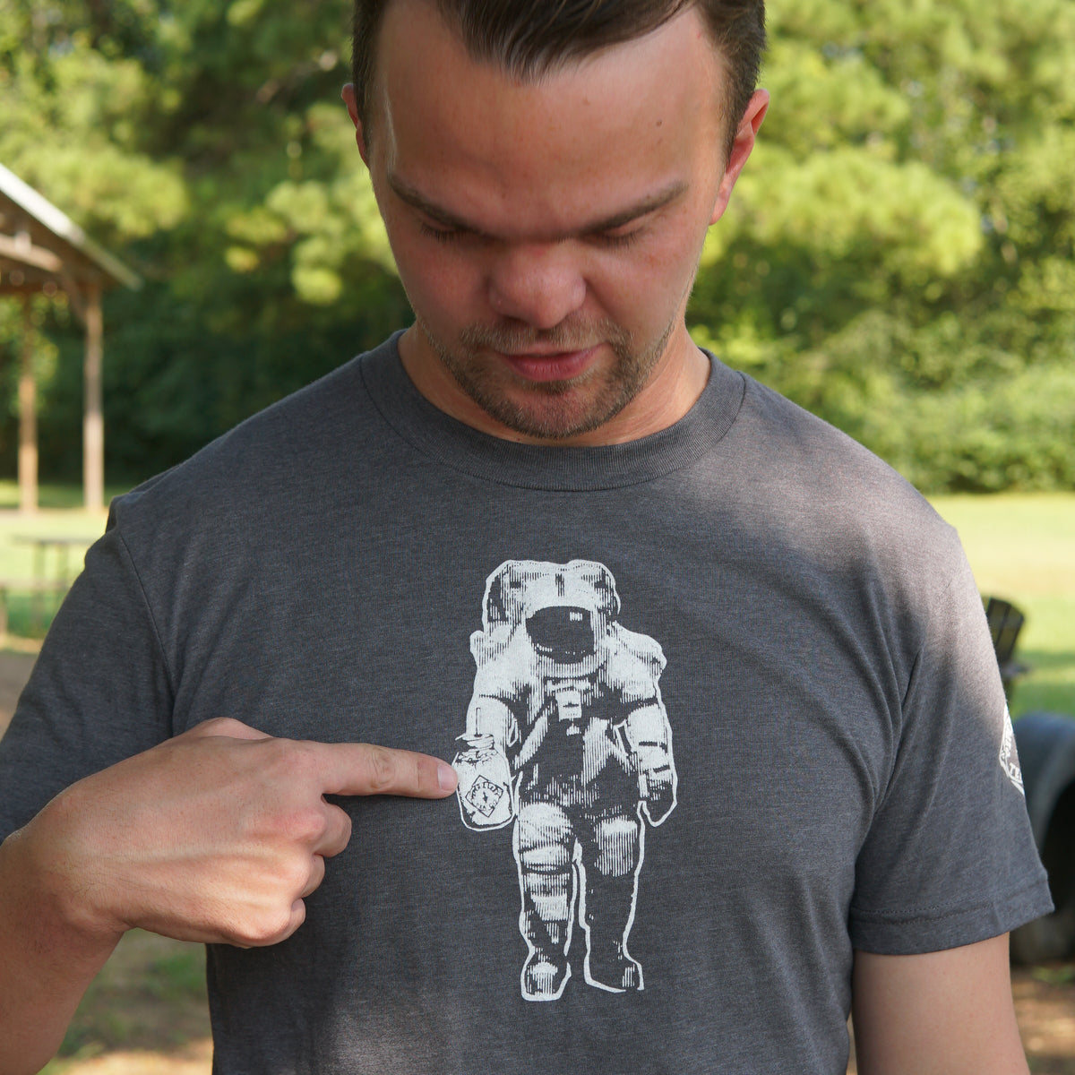 Wearing the Piper & Leaf Tea Co. Space Man Classic Tee-Shirt in grey, a person points to its astronaut graphic while standing outside among the trees, proudly showing off their Rocket City spirit.