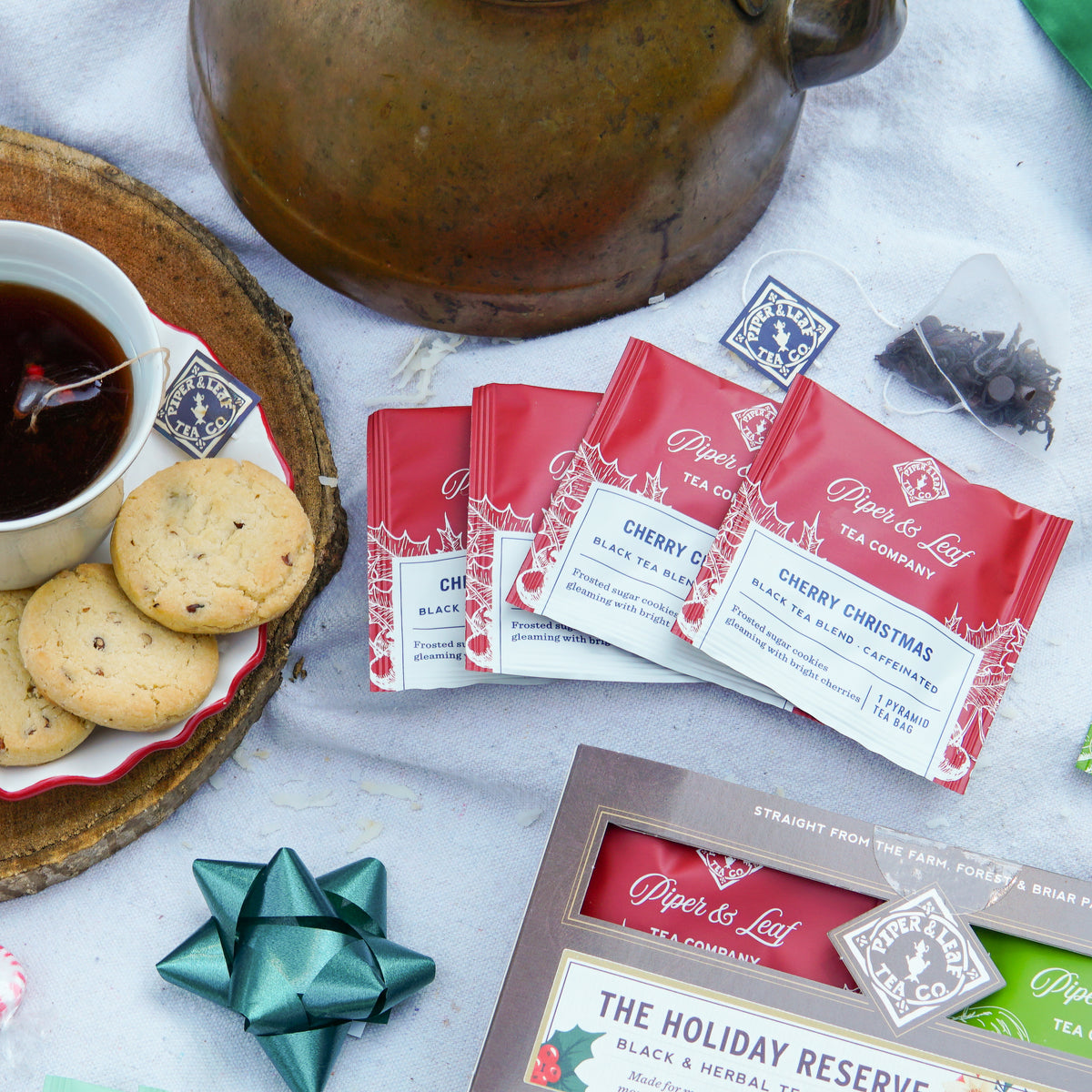 A cup of holiday tea with three cookies on a wooden coaster, beside four individually wrapped tea bags and a box of Holiday Reserve - Box of 16 Tea Bags by Piper & Leaf Tea Co.