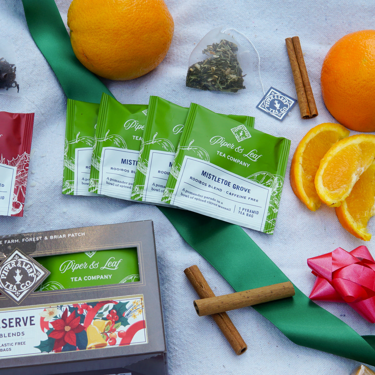 A Piper & Leaf Tea Co. Holiday Reserve - Box of 16 Tea Bags, individual tea bags, a loose rooibos infusion sachet, fresh orange slices, and cinnamon sticks arranged on a white cloth with a green ribbon.