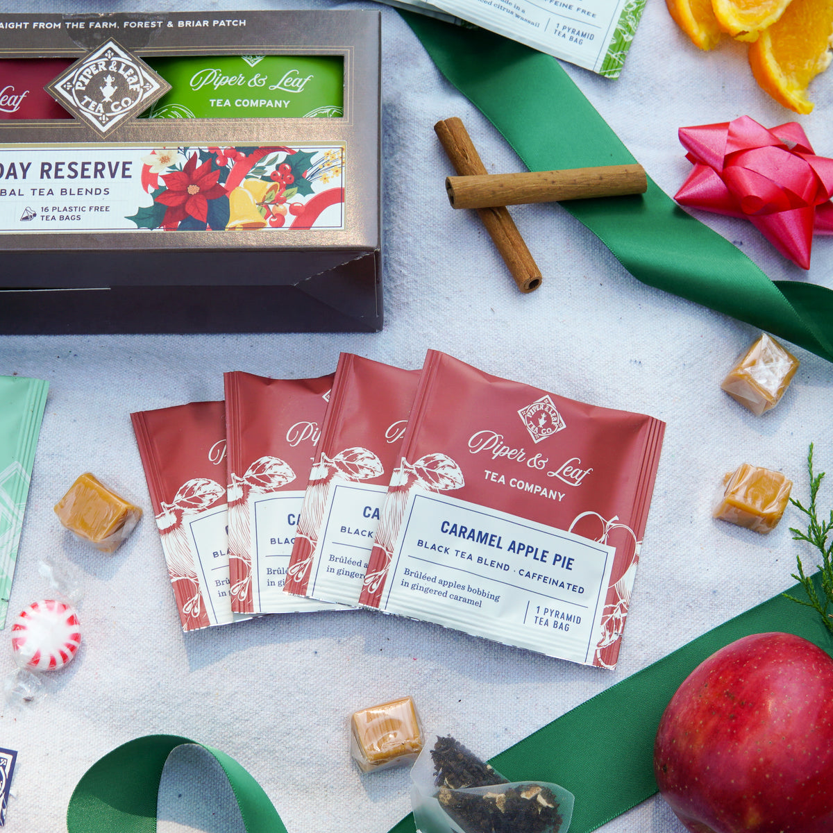 A Holiday Reserve - Box of 16 Tea Bags featuring four Caramel Apple Pie sachets, cinnamon sticks, caramel candies, a red apple, and a green ribbon, perfect for winter tea blends.