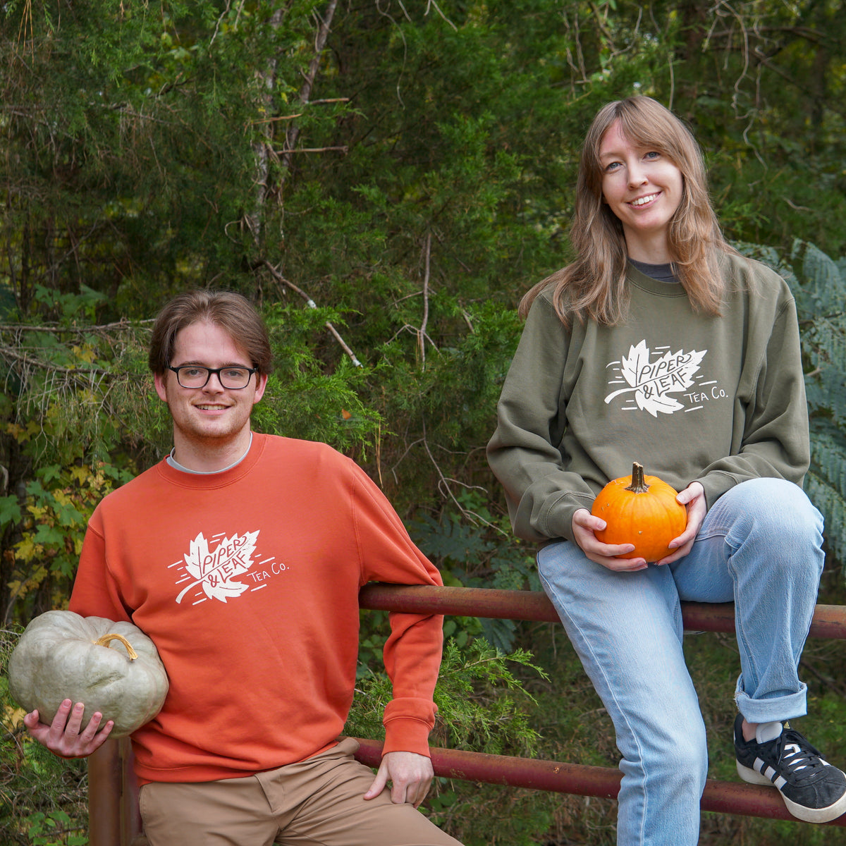 Guy in burnt orange Piper & Leaf tea co. sweatshirt outside leaning up against a fence where a girl is sitting on the fence wearing the same sweatshirt in an army green. 