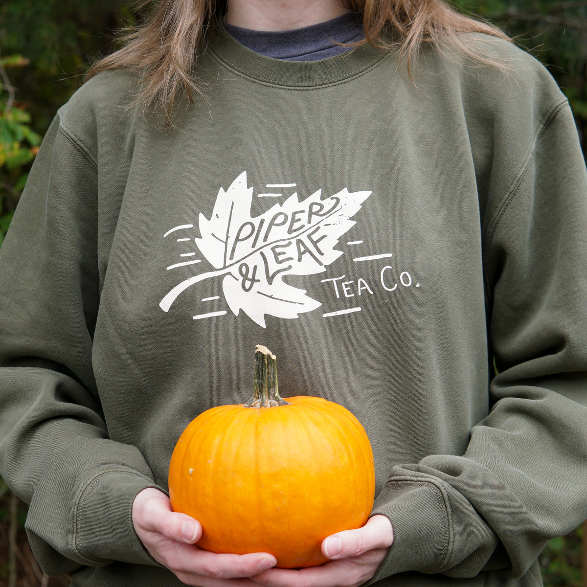 Girl holding a pumpkin while wearing a green PIper & Leaf Tea Co autumn leaf sweatshirt 
