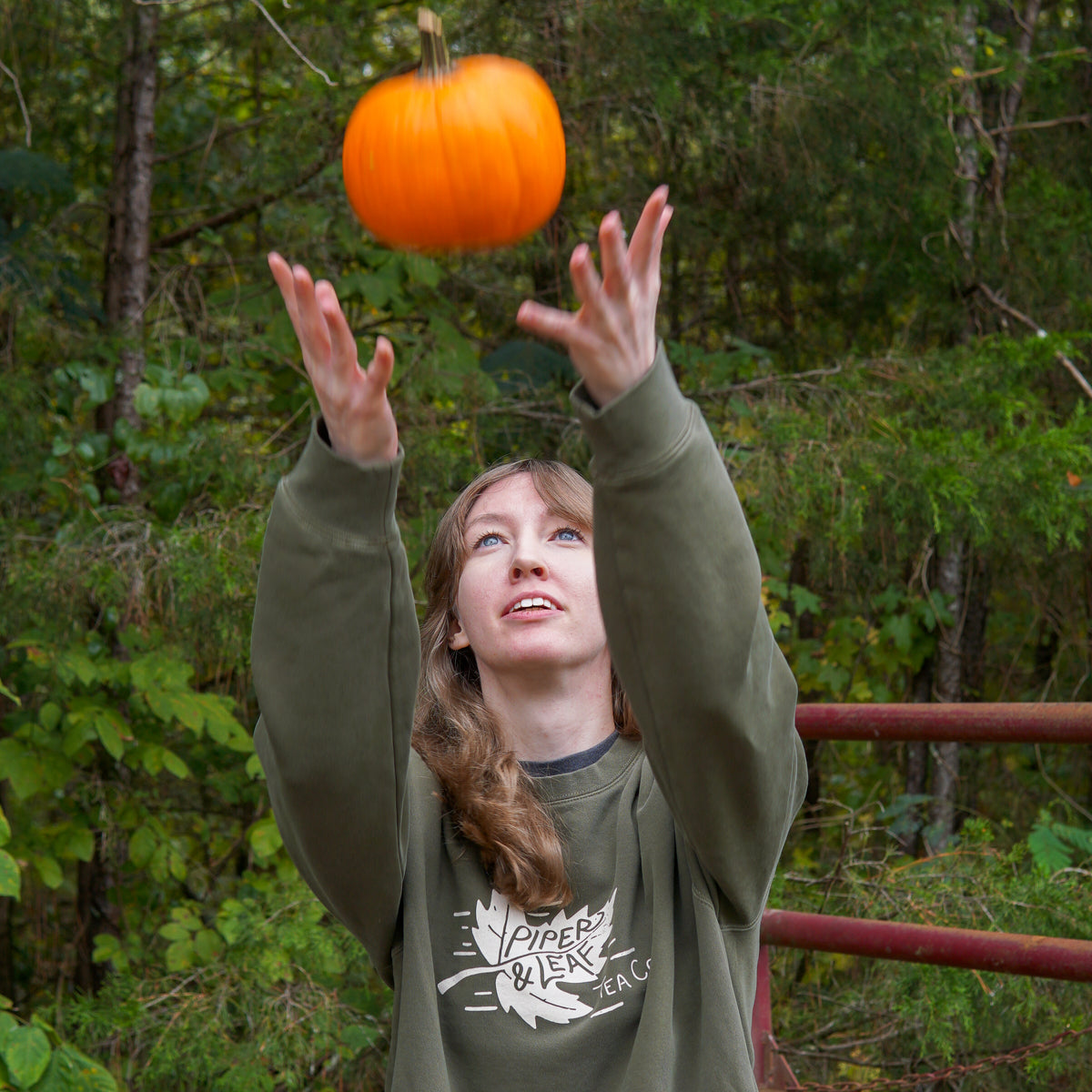 girl in green Piper & Leaf Tea co. autumn leaf sweatshirt throwing and catching a small pumpkin in the air. 