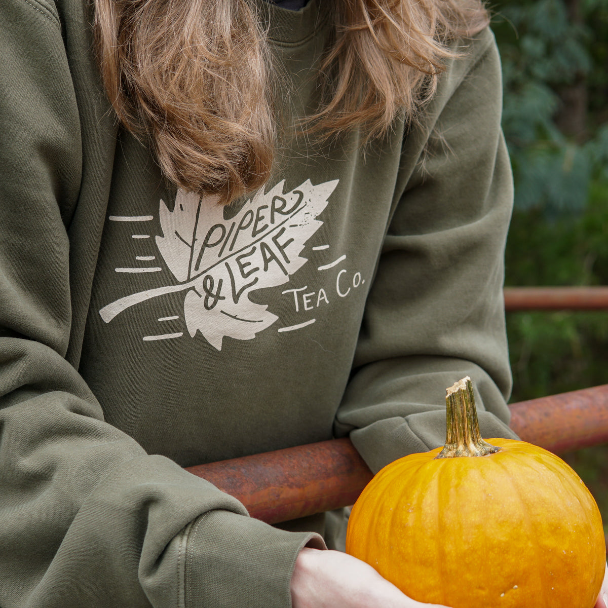 Girl leaning against a fence wearing a green  Piper & Leaf Tea Co. autumn leaf sweatshirt while holding small pumpkin. 
