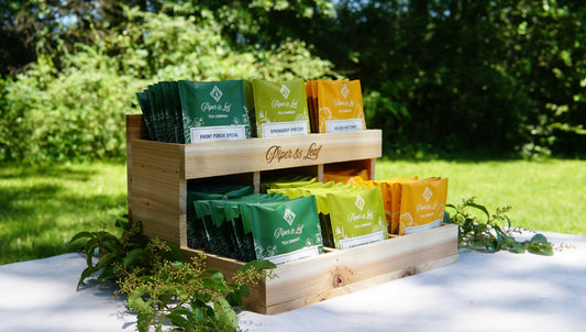 A Cedar Nook Display by Piper & Leaf Tea Co. holds assorted green, yellow, and orange tea bags outside on a white table with lush greenery—ideal for enjoying an al fresco tea tasting.