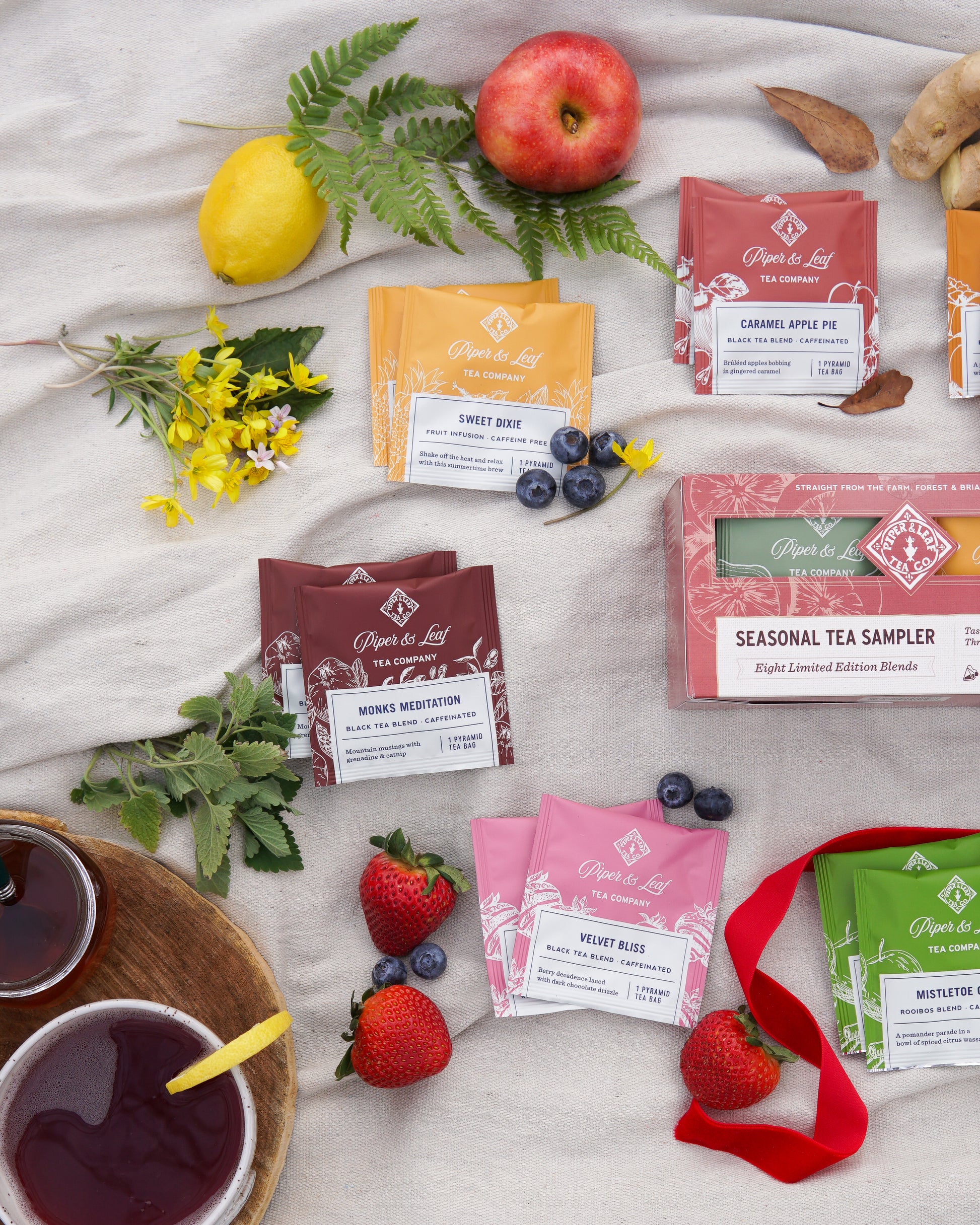 A Piper & Leaf Tea Co. Seasonal Tea Sampler (box of 16 tea bags), fresh fruits, a lemon, berries, wildflowers, and two cups of black tea are arranged on a light cloth background.