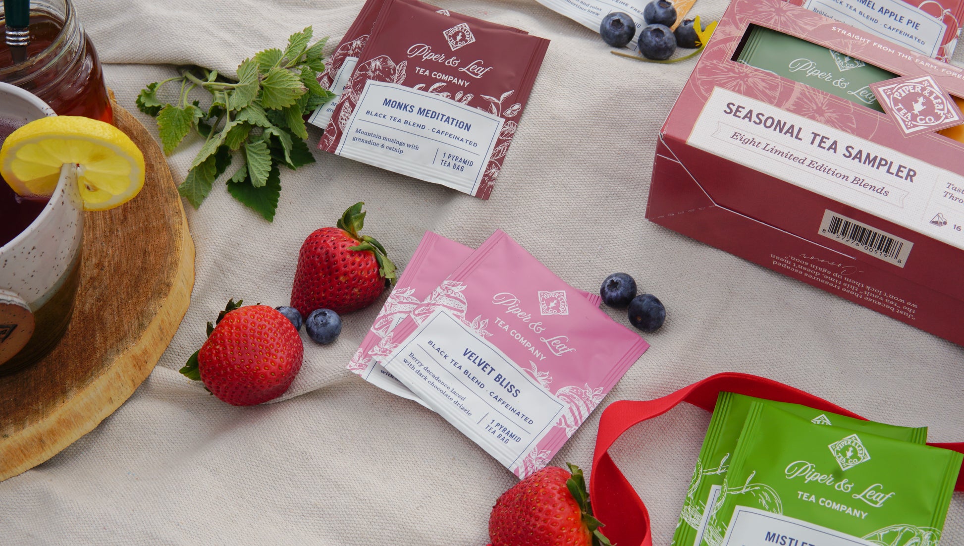 A Piper & Leaf Tea Co. Seasonal Tea Sampler (box of 16 tea bags) is arranged with fresh berries beside a glass of iced black tea garnished with lemon and mint, all set on a cloth surface.
