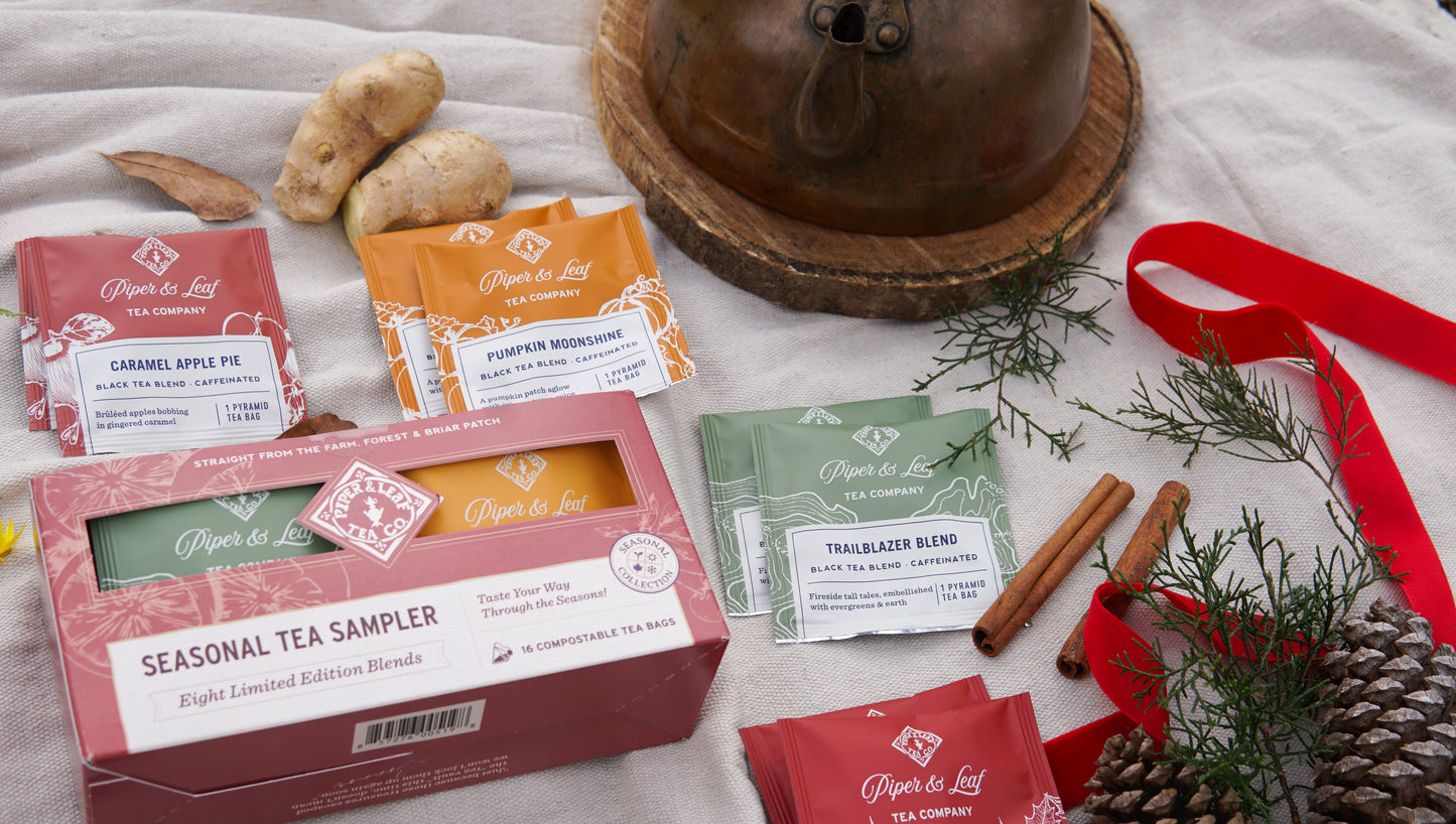 A Piper & Leaf Tea Co. Seasonal Tea Sampler—Box of 16 Tea Bags—is displayed alongside ginger, cinnamon sticks, pine, a copper kettle, and a red ribbon. Enjoy the perfect black tea blend for any season.