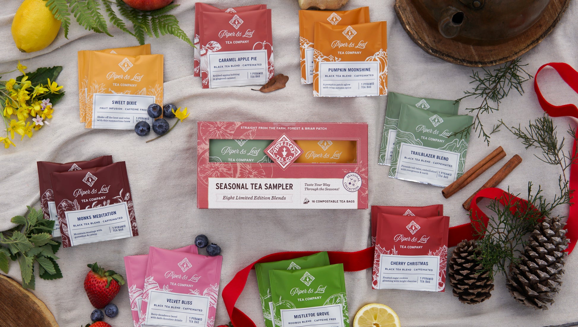 A Piper & Leaf Tea Co. Seasonal Tea Sampler—Box of 16 Tea Bags—is displayed on a table with assorted colorful tea packets, fruits, herbs, pinecones, and cinnamon sticks.