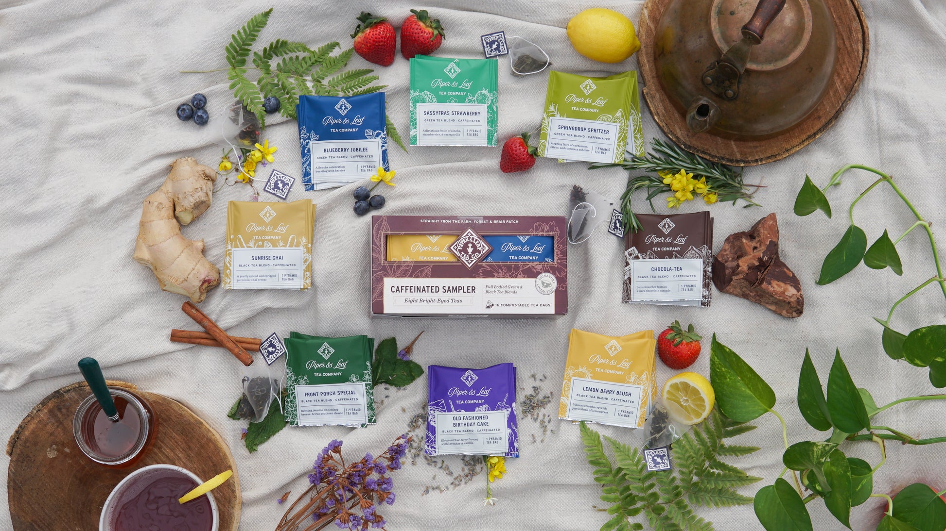 Piper & Leaf Tea Co.'s Caffeinated Sampler—Box of 16 Tea Bags—displayed on a cloth with fresh fruits, herbs, a teapot, a cup of green tea blend, flowers, and spices.