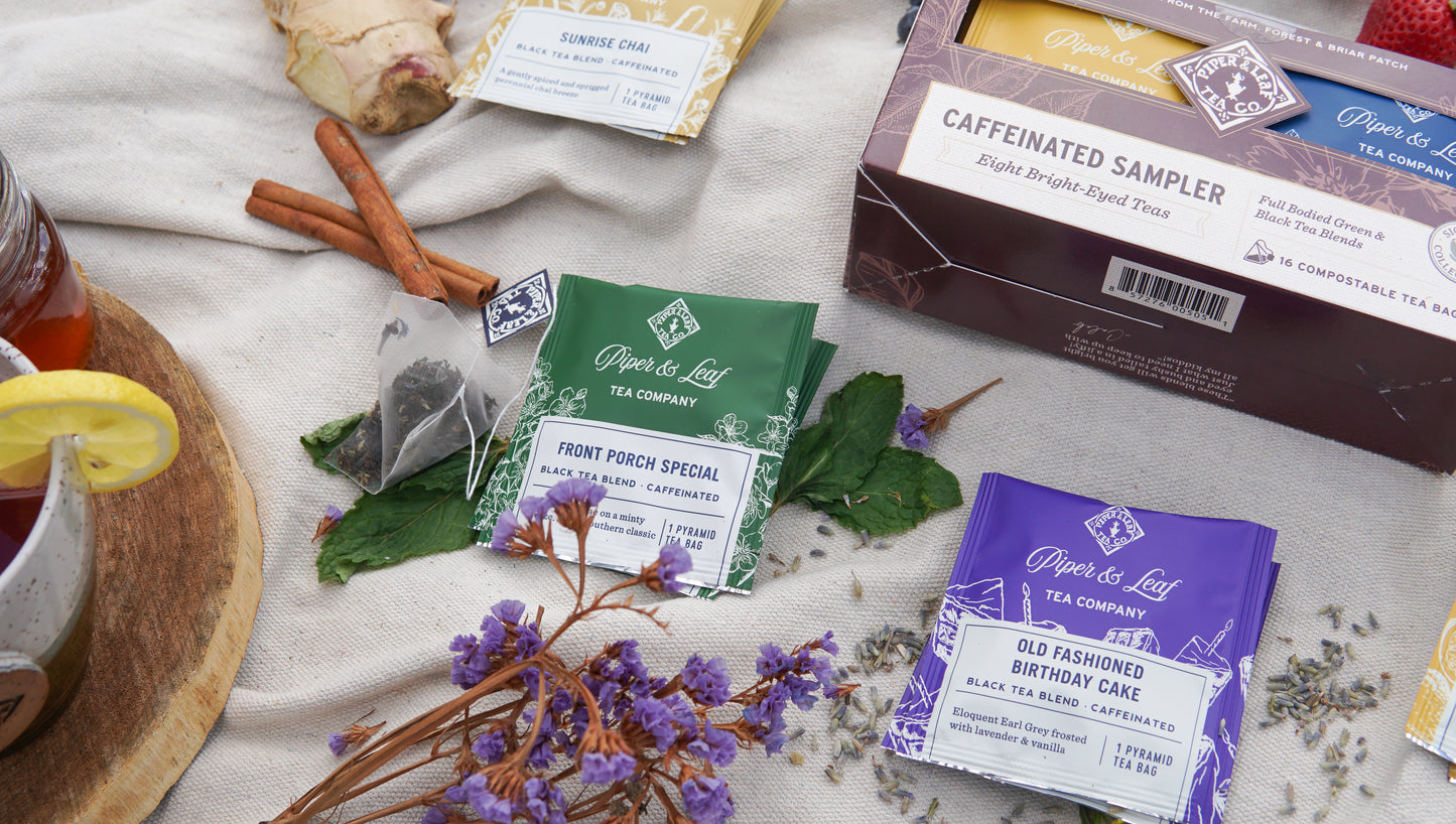 Assorted tea packets, a tea bag, cinnamon sticks, and lavender flowers rest on a beige cloth beside the Piper & Leaf Tea Co. Caffeinated Sampler – Box of 16 Tea Bags in the background.