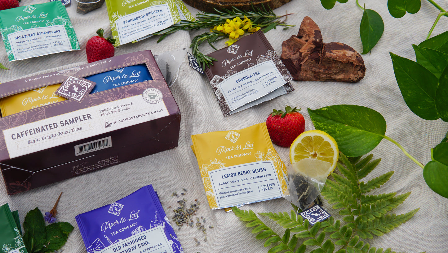 The Piper & Leaf Tea Co. Caffeinated Sampler—Box of 16 Tea Bags—featuring green and other caffeinated blends, is displayed on cloth alongside strawberries, lemon slices, ferns, and herbs.