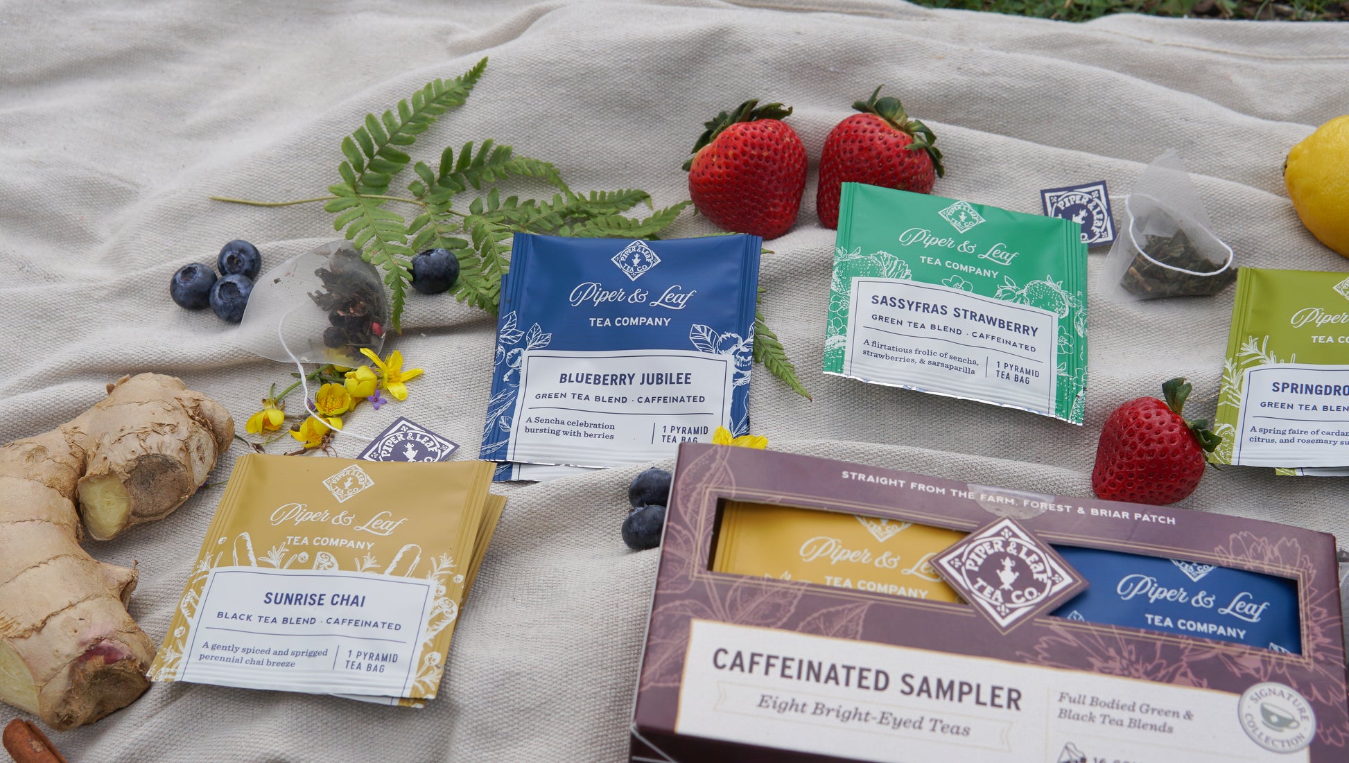 Piper & Leaf Tea Co.’s Caffeinated Sampler - Box of 16 Tea Bags is displayed on cloth with fresh berries, ginger, tea bags, and flowers.