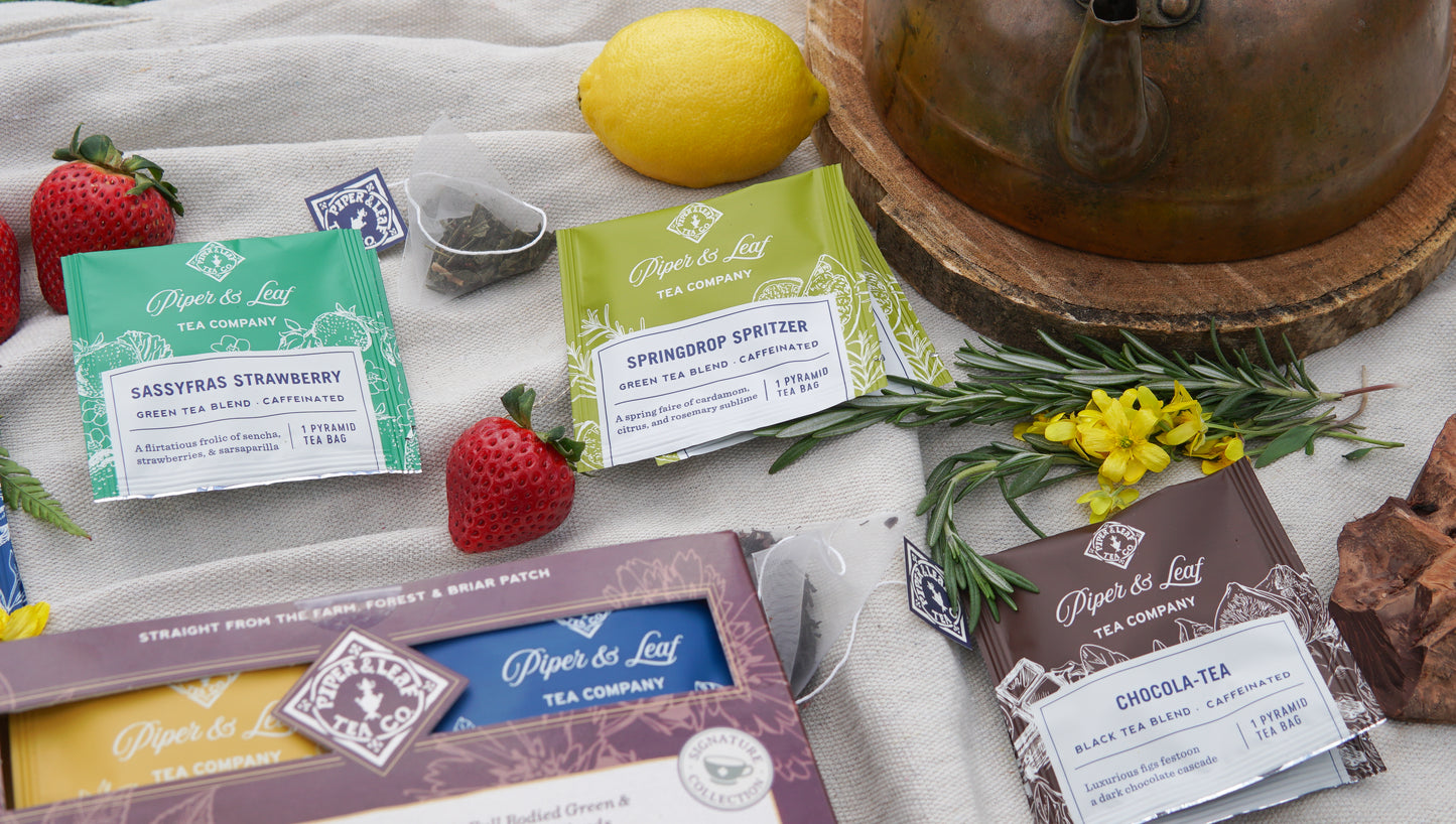 Boxes of Piper & Leaf Tea Co.'s Caffeinated Sampler—16 tea bags in assorted flavors—are displayed on a table with fresh strawberries, a lemon, herbs, and a copper kettle.