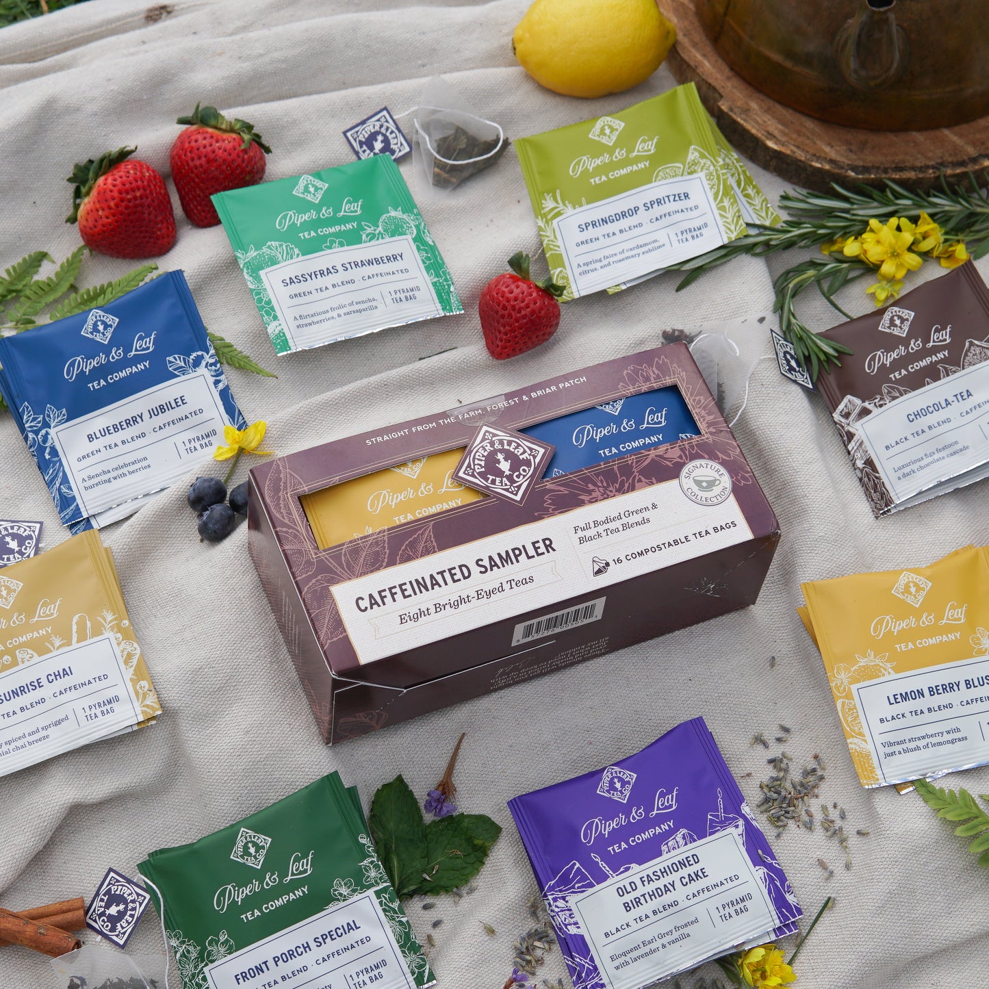 Piper & Leaf Tea Co.’s Caffeinated Sampler—Box of 16 Tea Bags—is displayed outdoors with assorted tea packets, including green tea blends and fresh fruits, herbs, and flowers arranged on a light cloth.