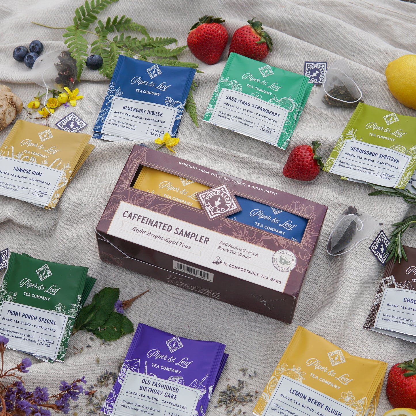 A box of Piper & Leaf Tea Co. Caffeinated Sampler—16 tea bags—rests on a cloth, surrounded by colorful caffeinated blends, fresh fruits, and flowers.