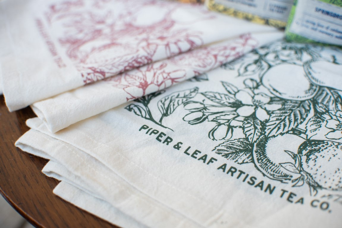 Tea towels featuring decorative prints of flora and fruit designs, one in green and one in red, proudly displaying “Piper & Leaf Tea Co.” on a wooden surface. Ideal as a charming housewarming gift or paired with the Mulling Spices Brewing Kit for a cozy touch.