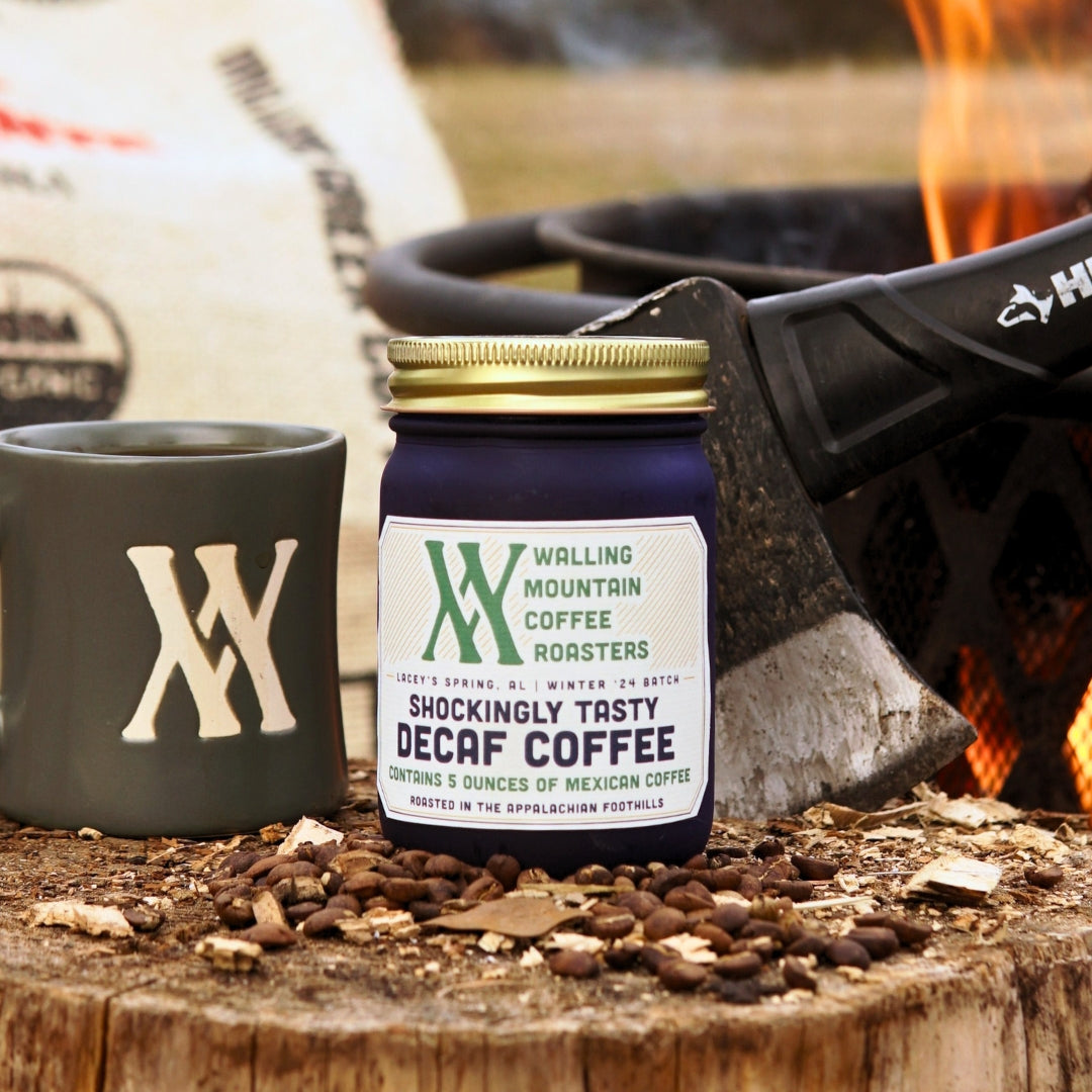 A 5oz jar of Walling Mtn. Decaf Coffee titled "Shockingly Tasty" sits by a matching mug on a log, with an axe and firepit nearby. This naturally decaffeinated Colombian coffee offers a medium dark roast without the buzz, ideal for cozy campfire moments.