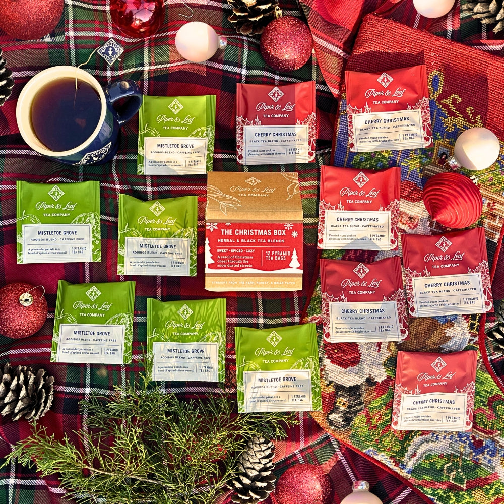 A holiday-themed arrangement featuring the "Christmas Box: Box of 12 Tea Bags" by Piper & Leaf Tea Co., accompanied by a cup of tea and festive decorations on a plaid blanket.