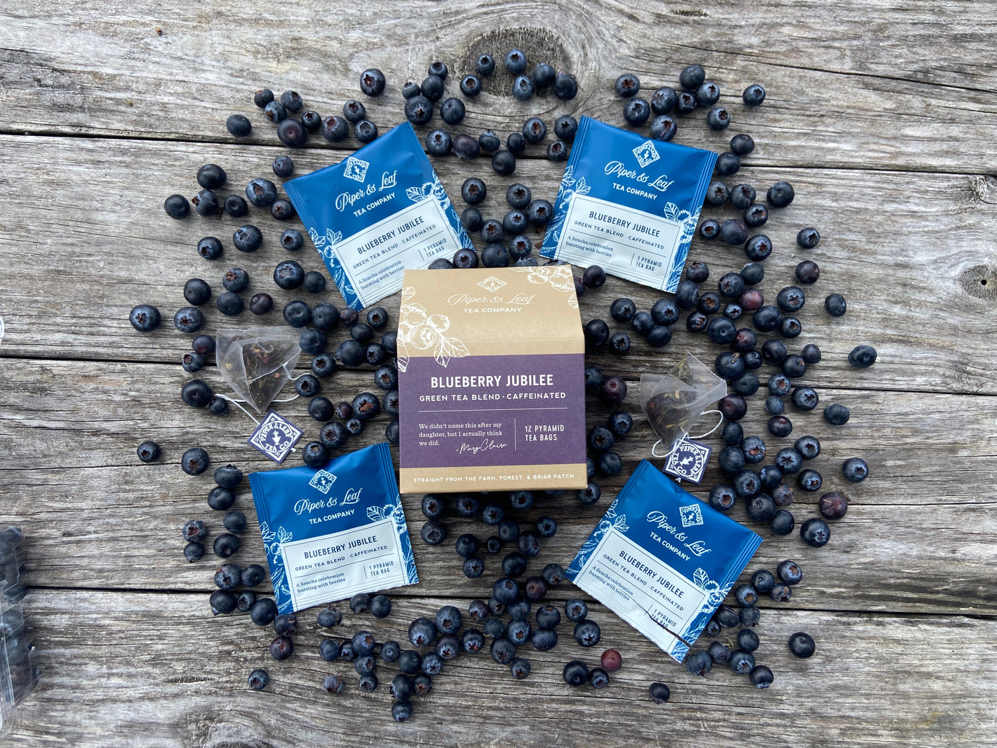 A box of Piper & Leaf Tea Co.'s Blueberry Jubilee—12 individually wrapped sachets—is displayed on a weathered wooden surface, surrounded by loose blueberries and five individual tea bags.