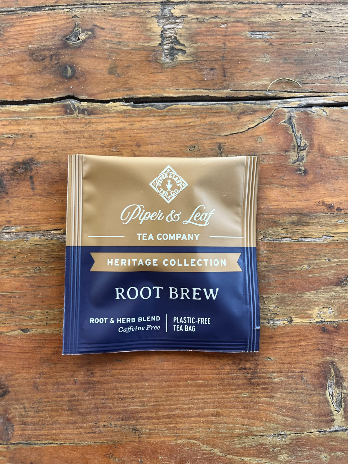 A Piper and Leaf Tea Co. “Root Brew” 50ct Individually Wrapped Tea Bag from the Heritage Collection rests on a wooden surface. The package is brown and navy, adorned with white and gold lettering.