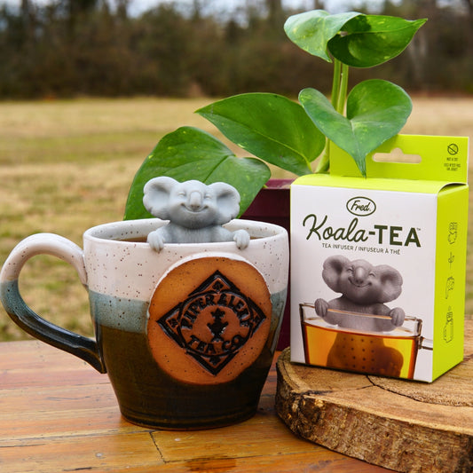 A Piper & Leaf Tea Co. ceramic mug sits on a table with a cute Koala-Tea Infuser beside a box labeled "Koala-Tea," making it an ideal koala-themed gift. A potted plant and grassy backdrop enhance the scene.