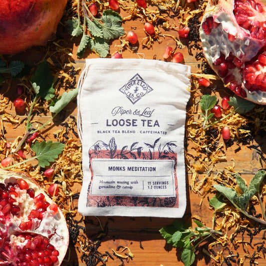 A bag of Piper & Leaf Tea Co.'s Monks Meditation Muslin Bag of Loose Leaf Tea, offering 15 servings, sits on a wooden surface amidst aromatic Ceylon Black Tea, vibrant pomegranate halves, and scattered leaves.