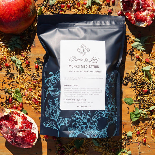 A "Monks Meditation" Pound Bag by Piper & Leaf Tea Co., offering 190 servings, is displayed with dried tea ingredients and pomegranate pieces on wood, highlighting its rich Ceylon Black Tea and grenadine flavors.