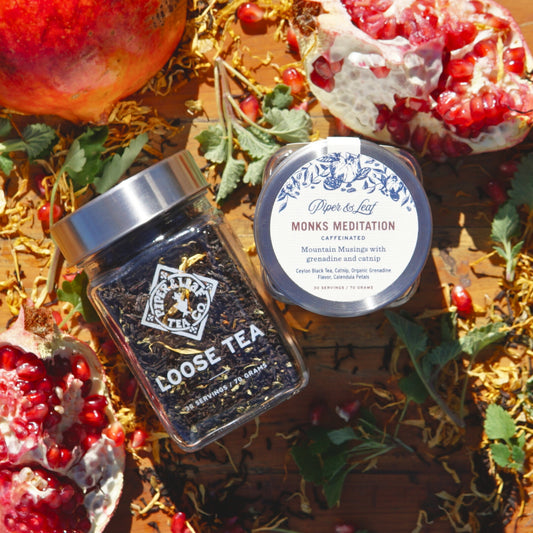 The Monks Meditation Glass Jar of Loose Leaf Tea by Piper & Leaf Tea Co., with its rich Organic Grenadine Flavor, sits alongside pomegranate halves and seeds on a wooden surface.