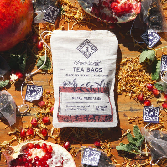 A "Monks Meditation 9ct Tea Bags in Muslin" package, from Piper & Leaf Tea Co., is elegantly surrounded by pomegranate seeds, dried leaves, and catnip on a wooden surface.